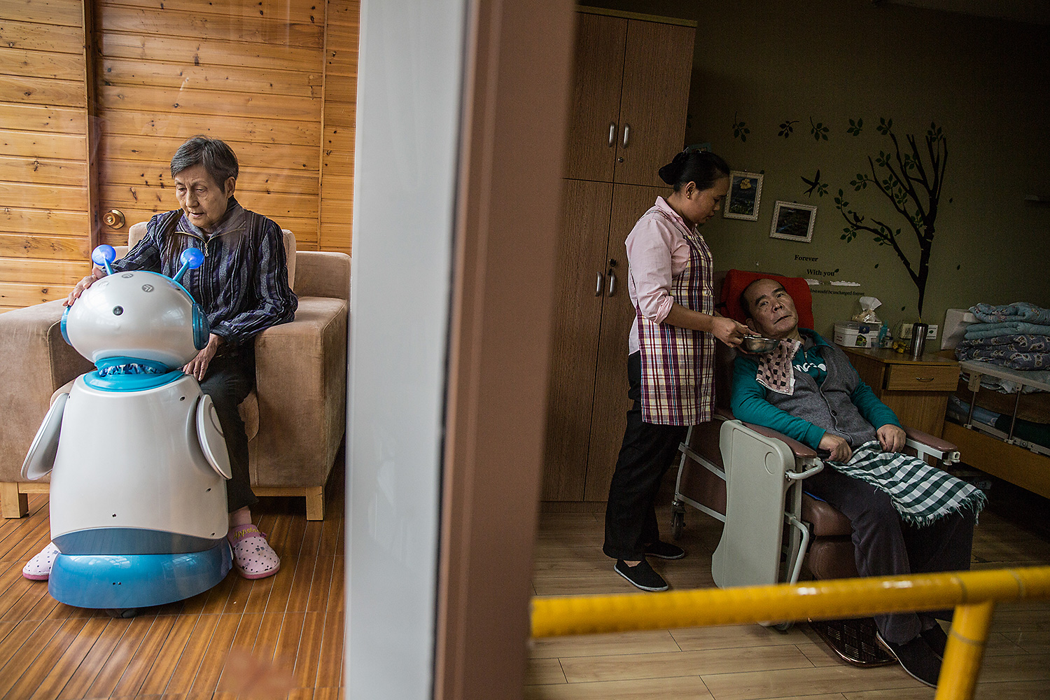 Zhao Qiuying, left, sits with an elder-care robot called Ah Tie, while a staff caregiver looks after an elderly man in an adjacent bedroom at the Hangzhou Social Welfare Center, Zhejiang province. Photo by Chen Ronghui @chenronghui1989