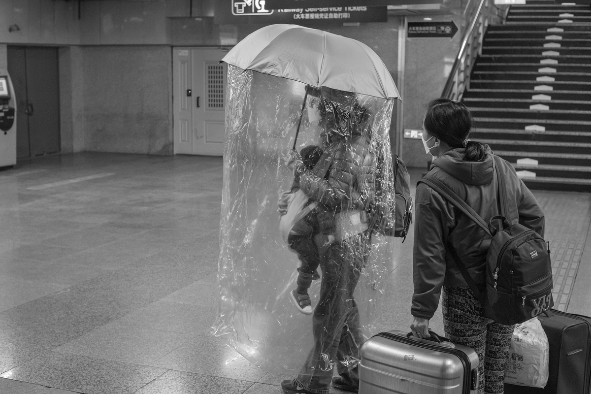 A parent carries an umbrella draped with a plastic sheet, enclosing them and their young child, Beijing West Railway Station, February 29, 2020.