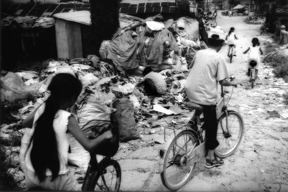 Children ride past computer components, imported from North America, Europe, and Japan. The parts, many of which are toxic, are dismantled and their recyclable materials extracted for sale in what was, less than a generation ago, a bucolic agricultural village outside of Guiyu, Guangdong.