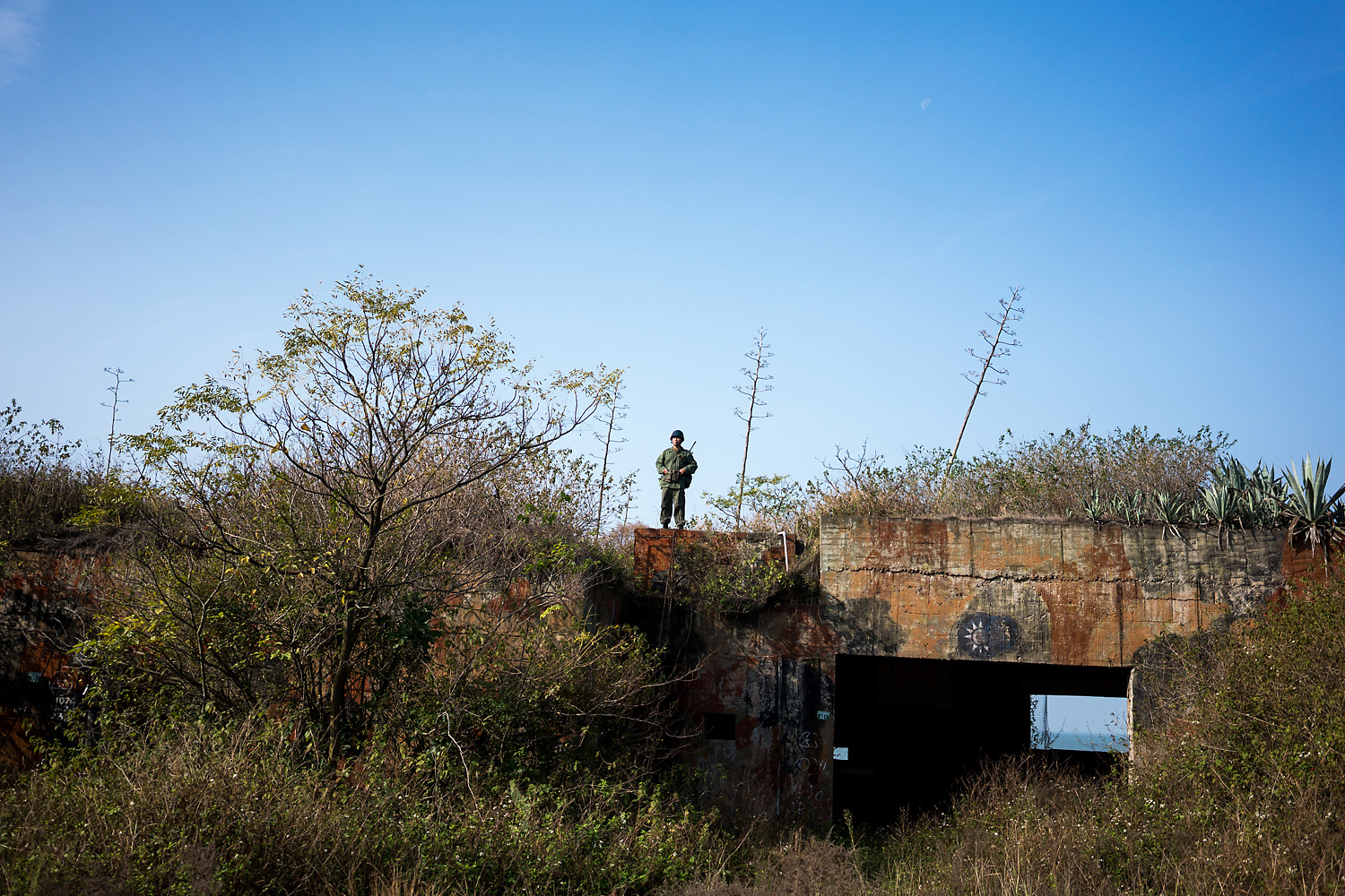 At the L18 military installation on Lieyu Island, overgrown with shrubs. Chen Zhixuan, dressed in army camouflage for a commemorative event, stands atop a bunker bearing the symbol of the KMT. On this day, a group of Kinmen residents gathered to explore the installation and clean the nearby beach of litter that had floated over from mainland China. The events were organized by the non-governmental organization Kinmen Battle History Association and activists in Kinmen’s burgeoning civil society scene to drum up support against the government’s plans to develop the installation into a tourist spot. Chen, a resident of Kinmen who works as a quality inspector in a cement factory, was wearing his military jacket,  circa the late-90s, when he served in the military on Kinmen. A few kilometers away, across the waters on Xiamen’s coast, stands a giant red banner that reads, ‘Peaceful reunification; One country, two systems.’